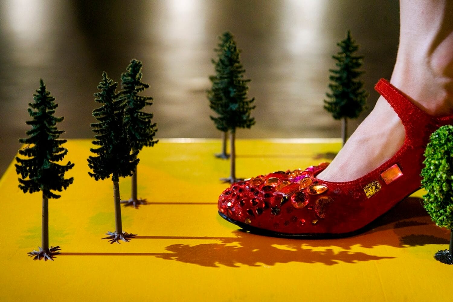 a close-up of a foot in a red shoe decorated with sparkling gems and sequinned embellishments, stepping on a yellow surface surrounded by miniature green fir trees resembling a small, crafted forest scene
