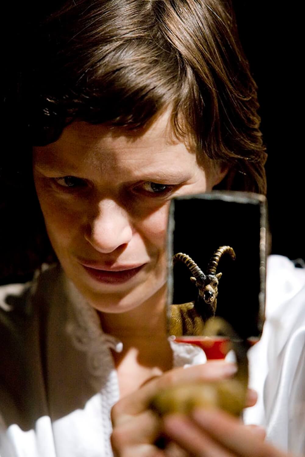 a person with long brown hair intensely gazing into a small mirror that reflects the image of a goat figurine with curved horns, creating a dramatic and mysterious composition