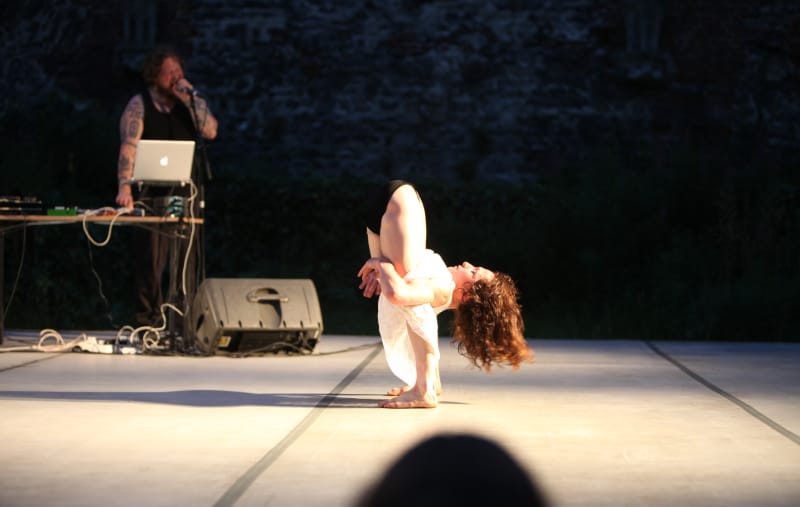 a contortionist stands on a side-lit stage with their head and body tucked right through their legs and out the other side, a voice artist and music technician stands at a computer in the background Iona Kewney © Tom Van Nuffel Purcell Room
