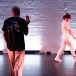 a performer on an indoor, back-lit stage, wearing a white casual costume, walks away from the audience along a brick wall, a second performers films them with a phone camera Birds show Kundle Cru