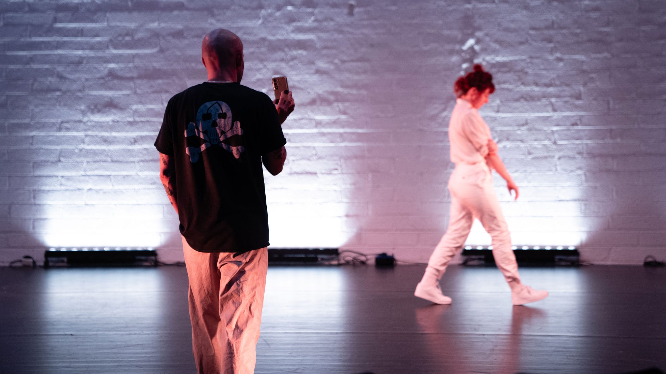 a performer on an indoor, back-lit stage, wearing a white casual costume, walks away from the audience along a brick wall, a second performers films them with a phone camera Birds show Kundle Cru
