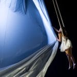 performer with long loose dark hair, wears a short floaty white sleeveless dress and bare feet in an indoor darkened stage, they are suspended mid-air by ropes and they swirl a large amount of fabric creating a sail under a soft blue light