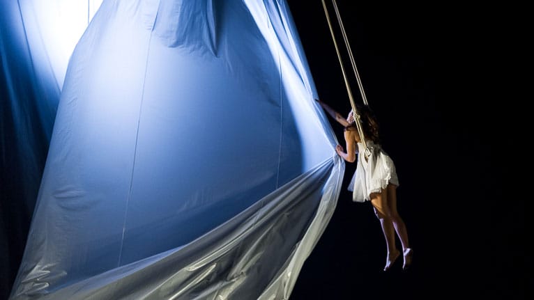 performer with long loose dark hair, wears a short floaty white sleeveless dress and bare feet in an indoor darkened stage, they are suspended mid-air by ropes and they swirl a large amount of fabric creating a sail under a soft blue light