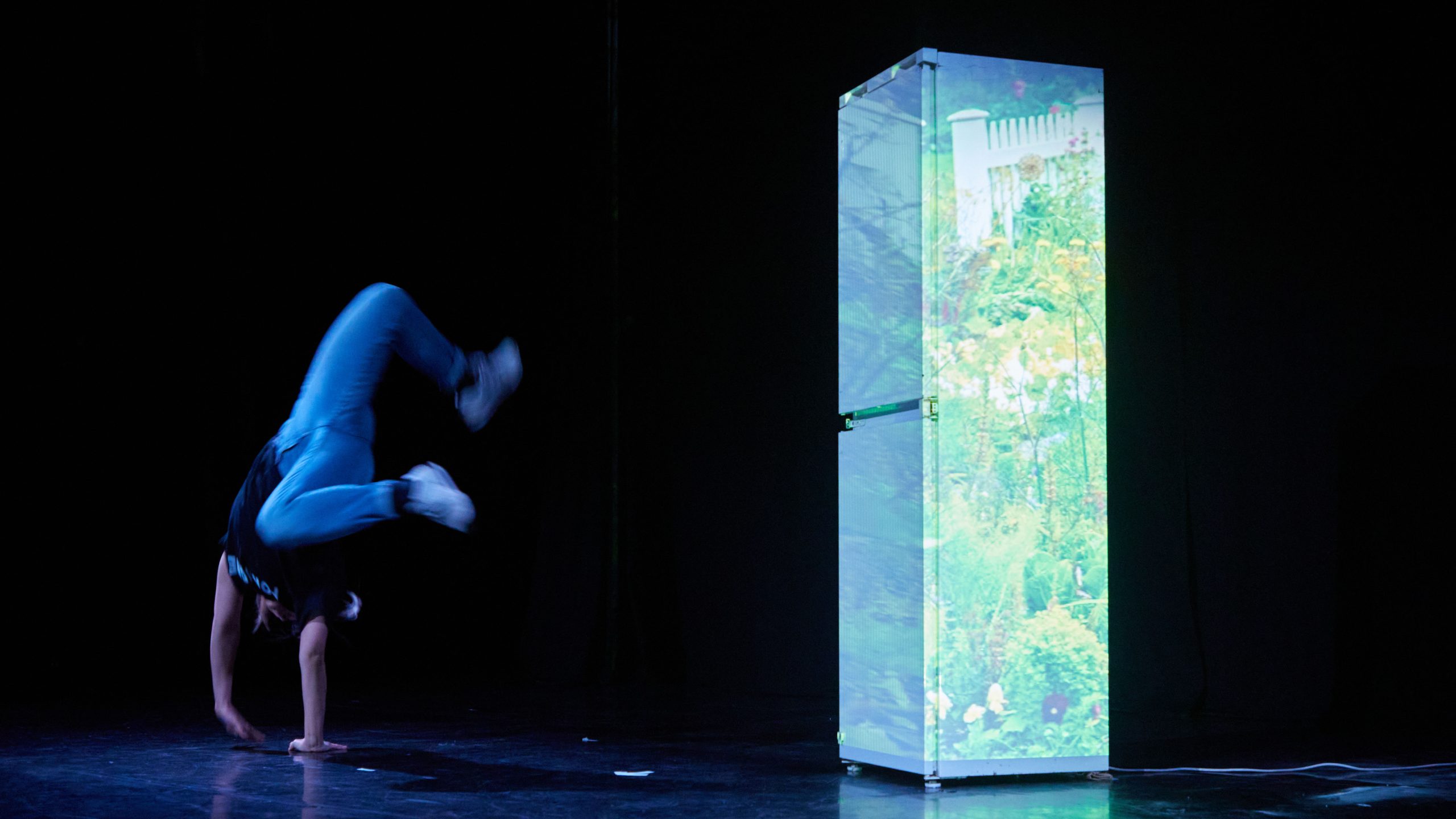 An east Asian non-binary performer, with a black t-shirt, blue jeans and white trainers in an indoor rehearsal space with a tall fridge behind them, which has a a garden scene projected on it, the performer is cartwheeling