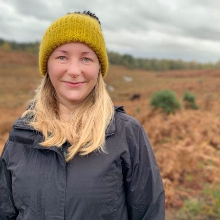 Lucy Babb has long blonde hair and wears a green wooden hat and grey waterproof jacket, behind is a dramatic grey sky and brown hillside outdoor scene