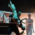 two dancers are spotlit on an indoor stage dancing against a washing line of cloths and sheets, behind the sheet there is a silhouette of a third performer