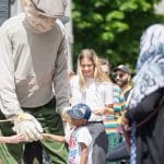 an outdoor audience engage with a giant puppet of an old man in a flat cap, the sun is shining and the audience are smiling