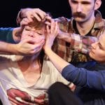 four dancers are wearing casual clothing on an indoor stage, three of them hold and push the head and face of the fourth performer