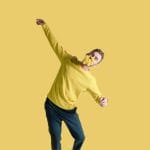 a dancer wearing dark trousers and a yellow top strikes a dance pose against a yellow background and with some daffodils in their mouth, they have their eyes closed and have short light brown hair