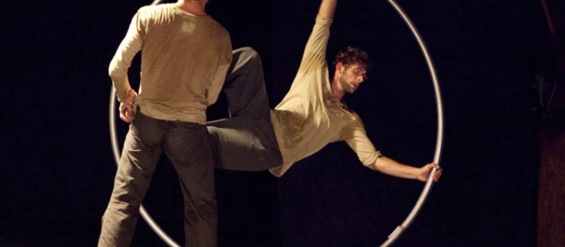 two acrobats both with brown hair and beards, wearing brown trousers and grey sweatshirts perform over a gym ball and with a cyr wheel in an indoor stage setting with yellow lighting widow stanton interview circoncentrique