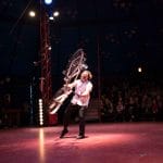 A performer spins a bicycle on stage, captivating the audience under a circus tent.