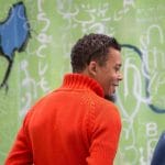 a Zimbabwean wearing a red polo neck sweater and smiling, stands slightly turned away against a green grafittied background, they have short dark curls and a goatee and wear glasses upon their head