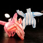 four coloured plastic bags on a dark stage are being blown around by a floor fan, it looks as though they are dancing