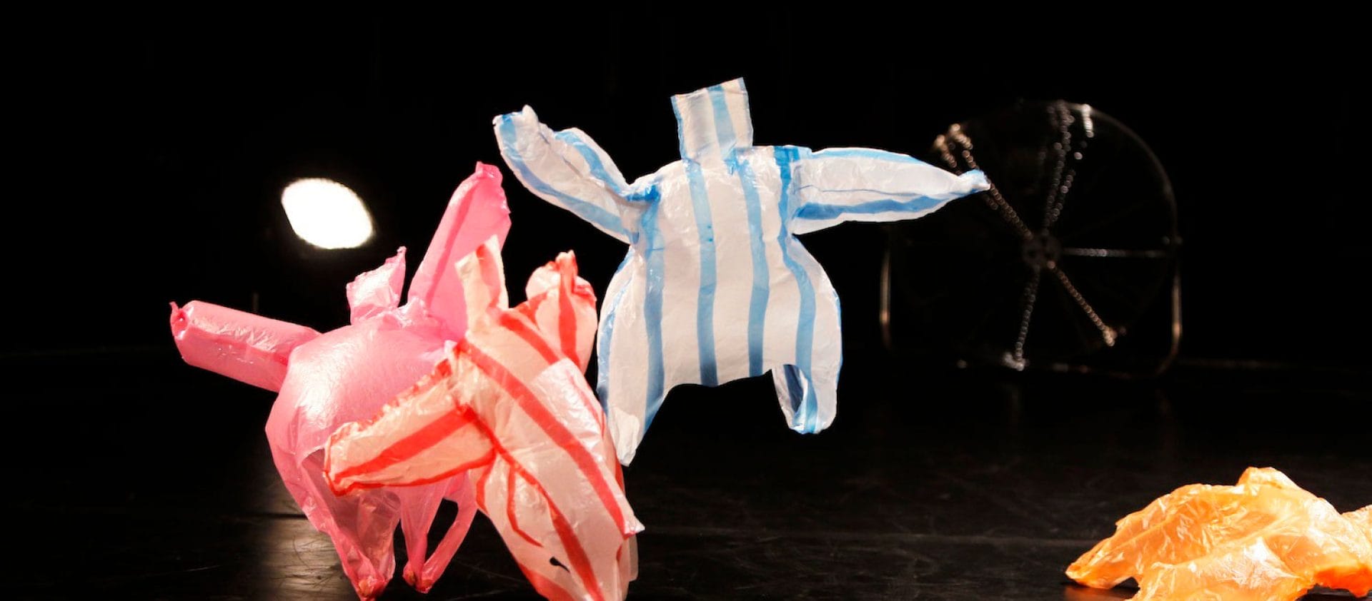 four coloured plastic bags on a dark stage are being blown around by a floor fan, it looks as though they are dancing