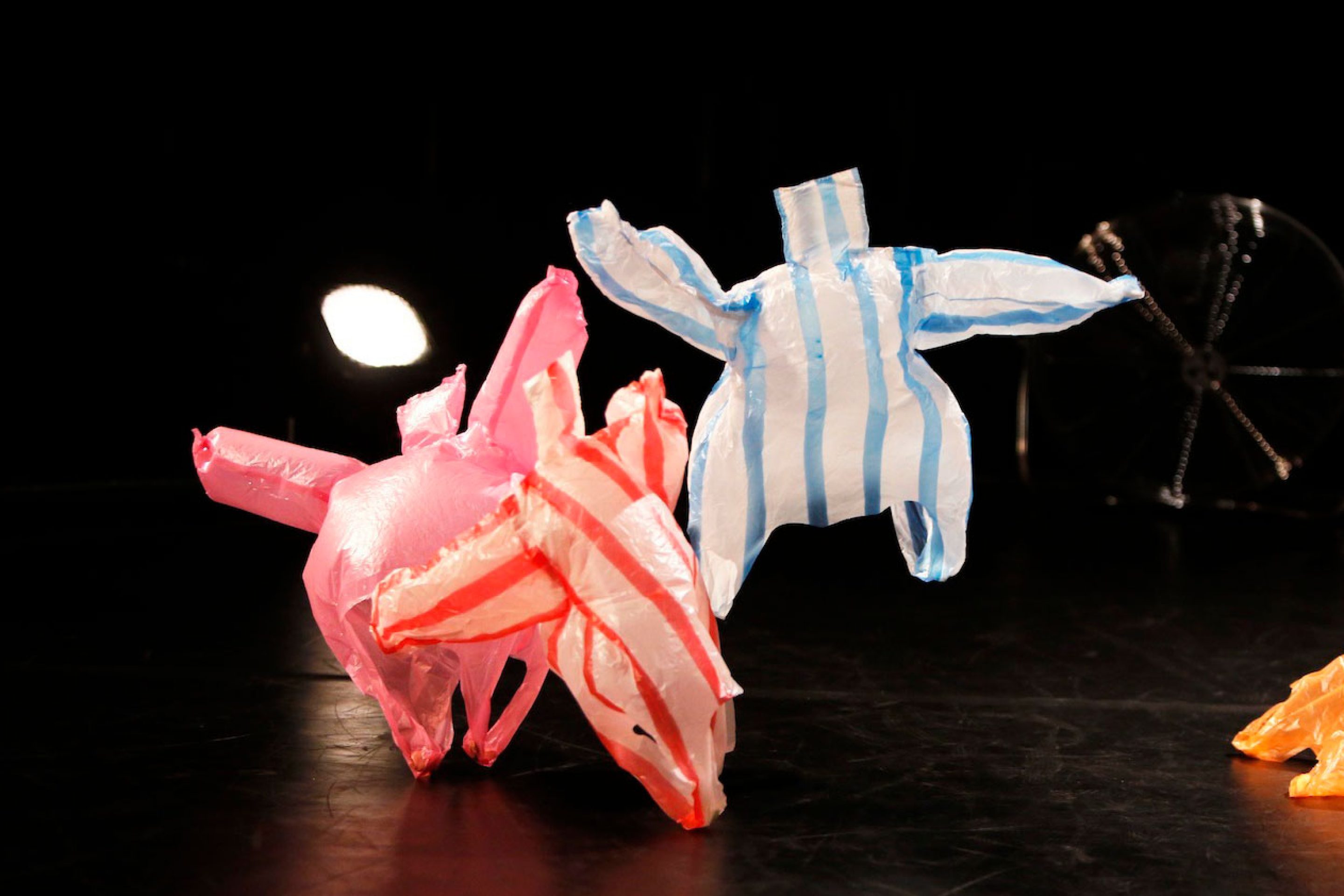 four coloured plastic bags on a dark stage are being blown around by a floor fan, it looks as though they are dancing