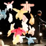 Many coloured plastic bags on a dark stage are being blown around by a floor fan, it looks as though they are dancing, one performer wearing black clothes and a hat holds one of the bags like a dance partner