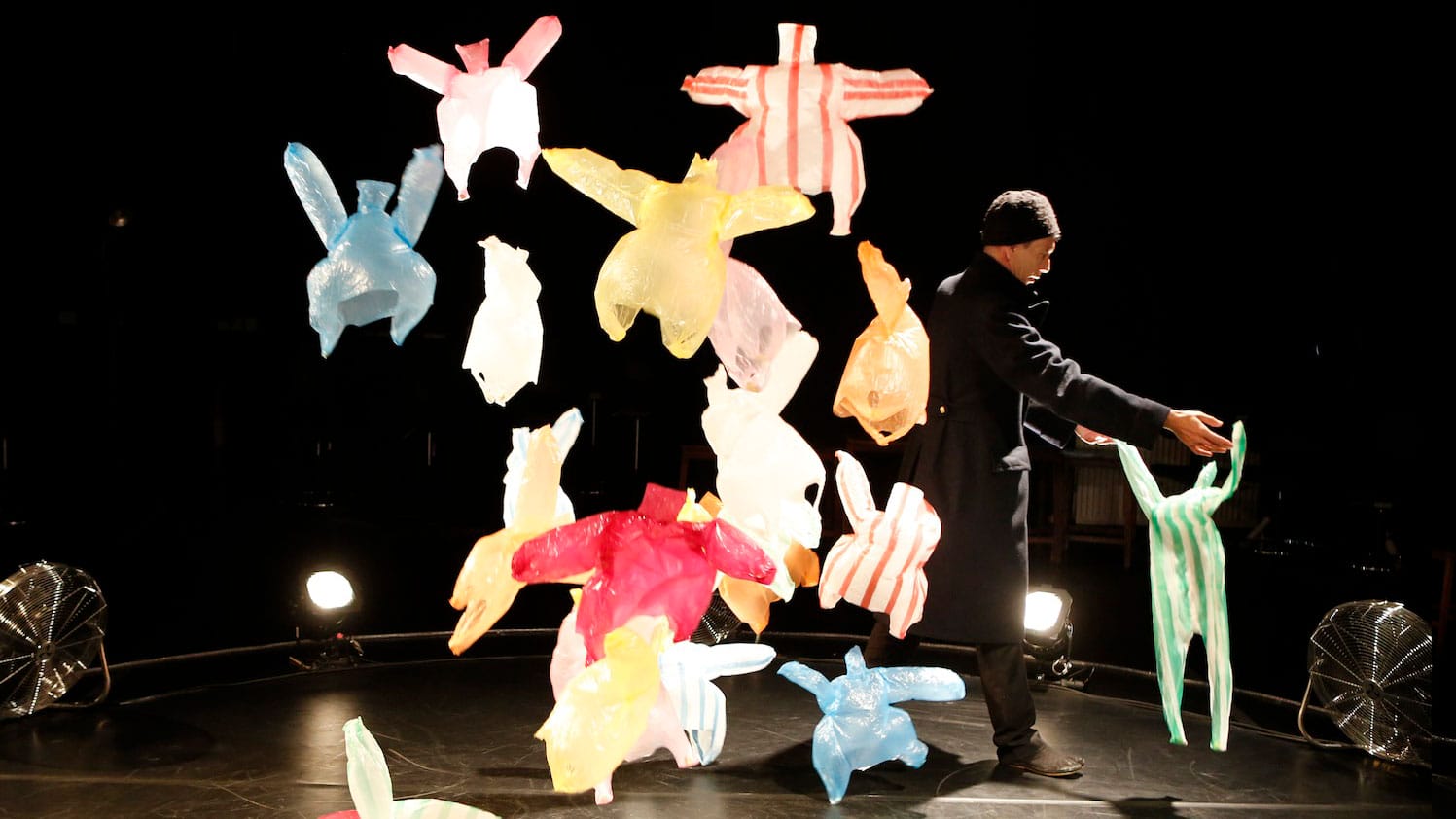 Many coloured plastic bags on a dark stage are being blown around by a floor fan, it looks as though they are dancing, one performer wearing black clothes and a hat holds one of the bags like a dance partner