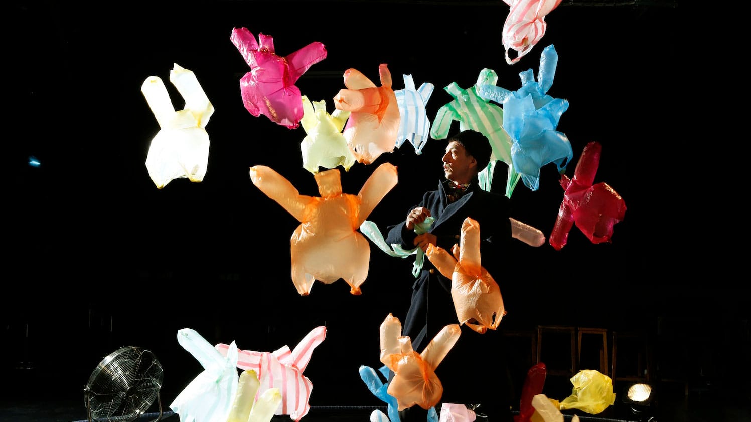 Many coloured plastic bags on a dark stage are being blown around by a floor fan, it looks as though they are dancing, one performer wearing black clothes and a hat stands in amongst the bags watching the action