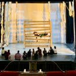 two dancers on a lit indoor stage climb through a giant wooden gate while eleven others sit and watch from the stage, the box seats in the theatre are either side of the image