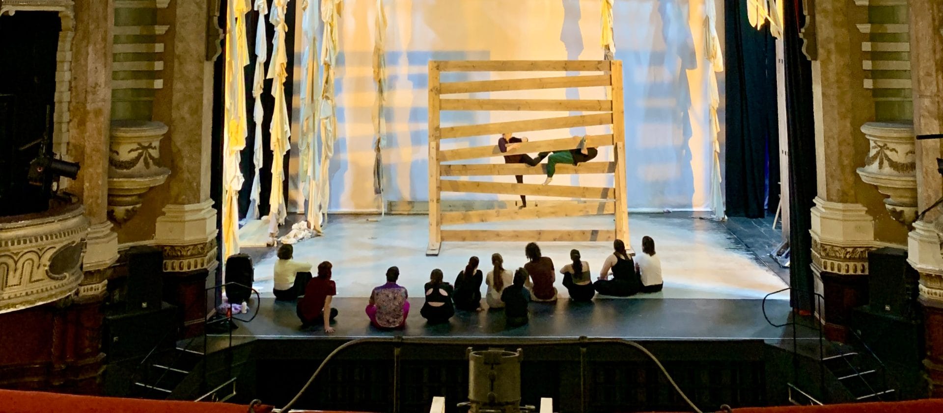two dancers on a lit indoor stage climb through a giant wooden gate while eleven others sit and watch from the stage, the box seats in the theatre are either side of the image