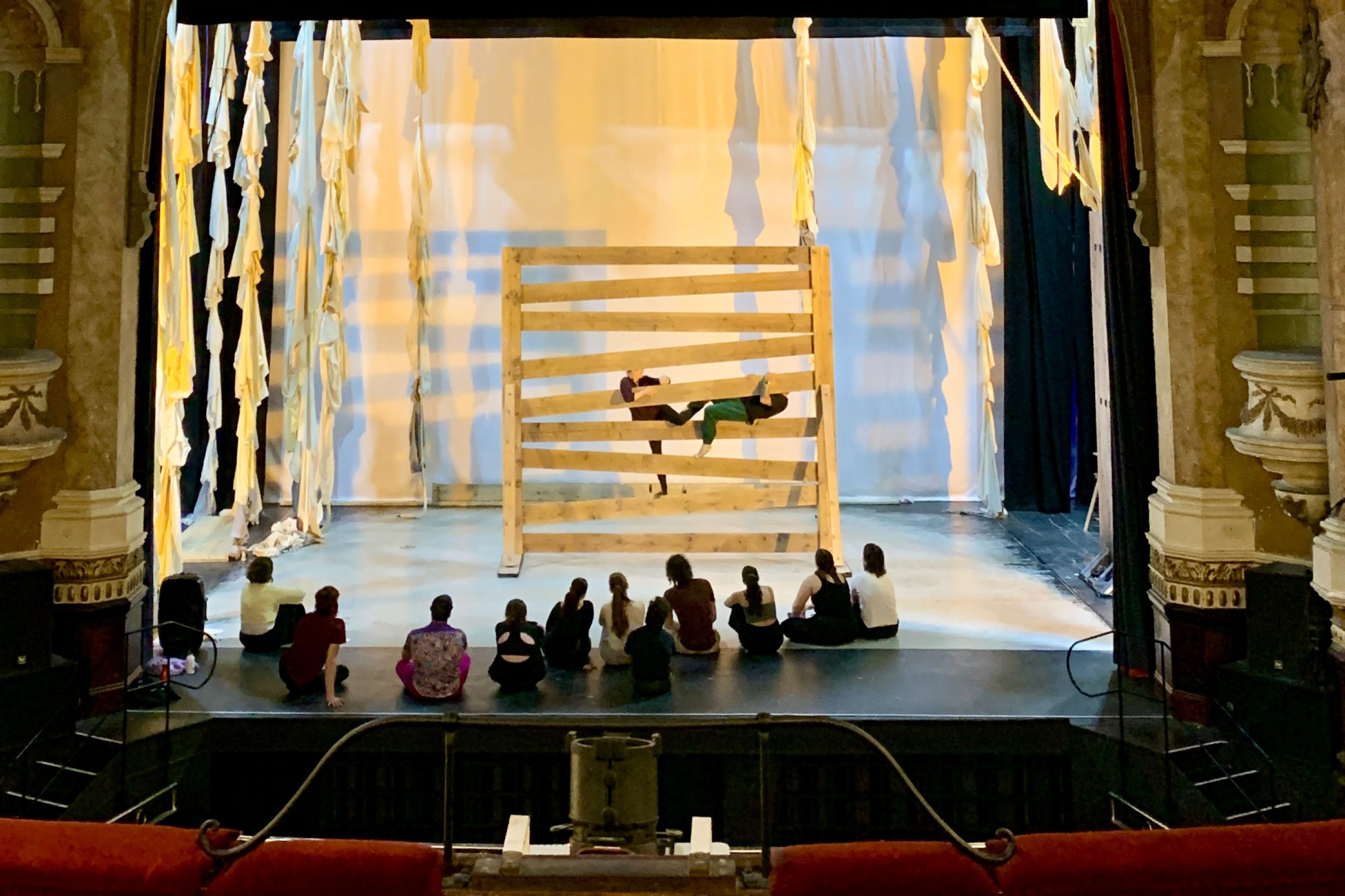two dancers on a lit indoor stage climb through a giant wooden gate while eleven others sit and watch from the stage, the box seats in the theatre are either side of the image