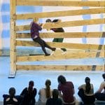 two dancers on a lit indoor stage climb through a giant wooden gate while eleven others sit and watch from the stage