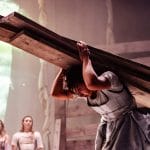 four actors on an indoor stage, three stand at the back looking at one performer in the foreground who bends while carrying four planks of wood on their back, they all wear pastel coloured dresses and aprons and have long fair hair, the event will be a masterclass in physical theatre