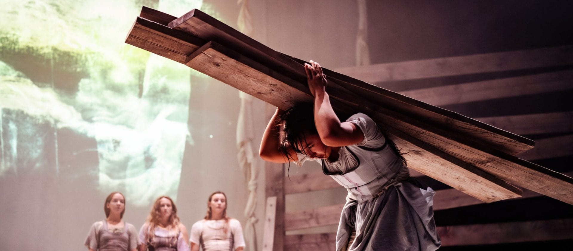 four actors on an indoor stage, three stand at the back looking at one performer in the foreground who bends while carrying four planks of wood on their back, they all wear pastel coloured dresses and aprons and have long fair hair, the event will be a masterclass in physical theatre