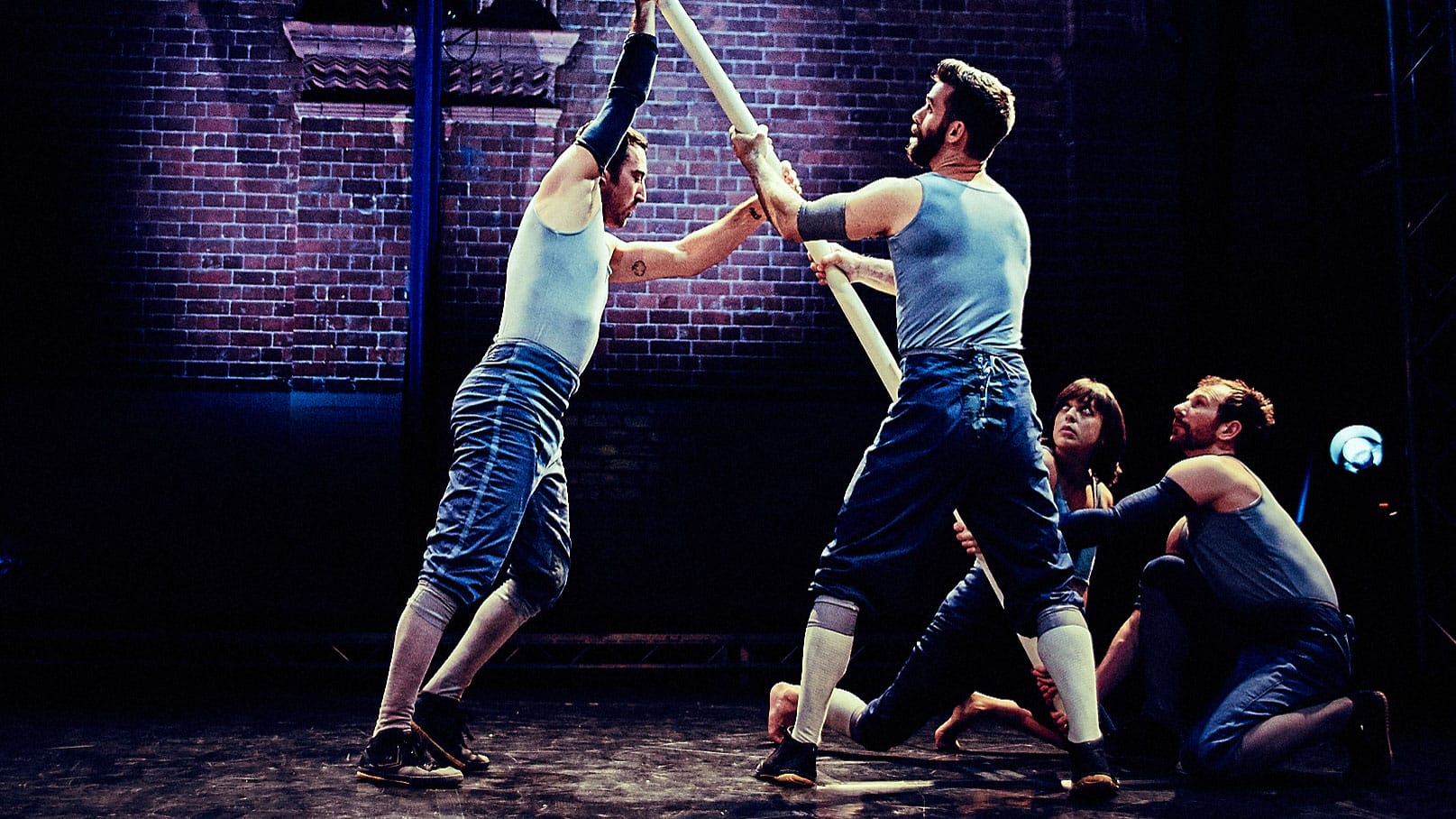 four performers hold a long scaffold pole between them creating a diagonal pole, they are on an indoor stage with a brick wall behind and are spotlit, all wear short blue jeans and pale blue vests