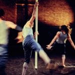 three performers move on an indoor stage with a brick wall behind and are spotlit, all wear short blue jeans and pale blue vests, one holds a long scaffold pole suspended from above but not touching the ground