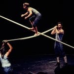 three performers hold two long scaffold poles between them creating a cross, a fourth performer climbs on the poles while a fifth watches, they are on an indoor stage and all wear short blue jeans and pale blue vests