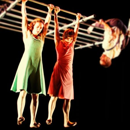 two performers with shoulder length hair, both in dresses, one red, one green and bare feet, hang by their hands from a criss cross of scaffold in a darkened indoor stage, they both look down and a third performer in a white shirt and dark trousers with braces hangs off the same equipment watching Circus Now: International Circus Exposure 2015 City Circ London network