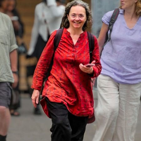 person walking across a bust road towards the camera wearing black trousers, a red shirt dress and a rucksack, they are smiling at the camera