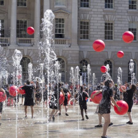 Performers juggle red balls and dance among fountains in front of a historic building. history of UK circus