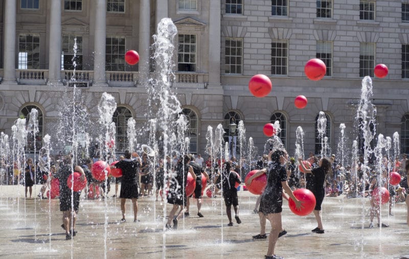 Performers juggle red balls and dance among fountains in front of a historic building. history of UK circus