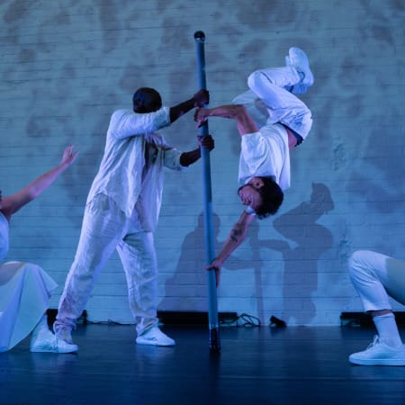 four dancers on an indoor, blue-lit stage, they all wear white casual costumes, two central performers hold a Chinese pole and perform on it, one upside-down, the other two performers kneel as they look on incl in summer 2024 activity round-up