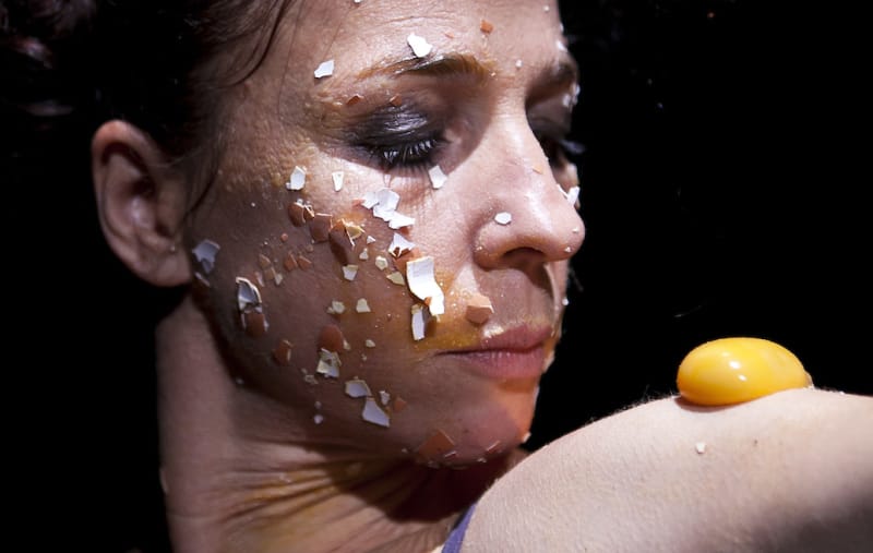 close up of a performer's face, with dark hair tied up and dark eye make-up, they have bits of egg shell stuck to their face and are looking intently at their own shoulder which has an egg yolk resting on it Jeanne Mordoj