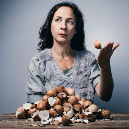 a performer wearing a fur and cloth grey short-sleeved top sits at a table looking off camera, on the table is a large pile of broken egg shells