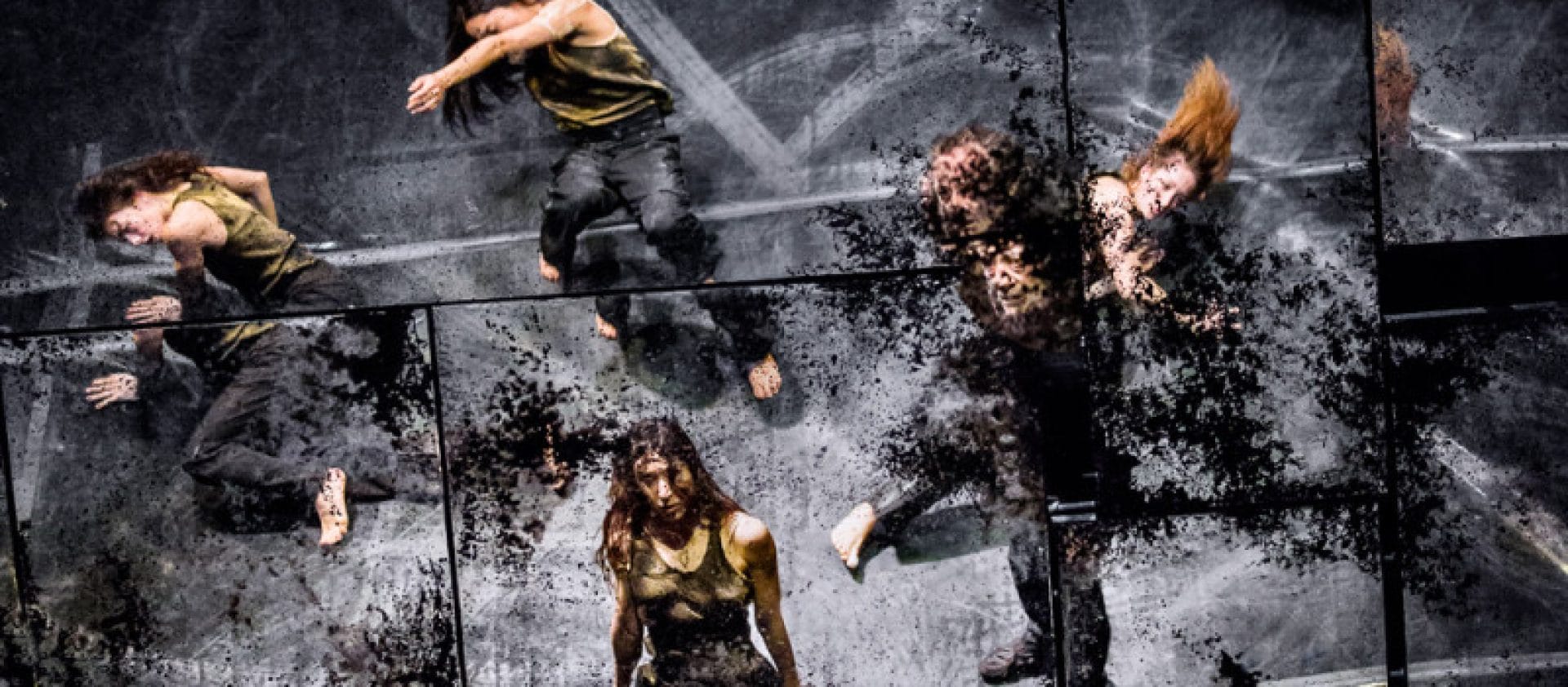 Performers stand on an indoor stage, dressed in a dark trousers and grey shirt and softly lit from above Performer stands on a messy desk on an indoor stage, dressed in a dark trousers and grey shirt and looks at the camera, surrounded by other performers dressed the same and softly lit by golden desk lamps James Thierrée Tabac Rouge Olivier Award