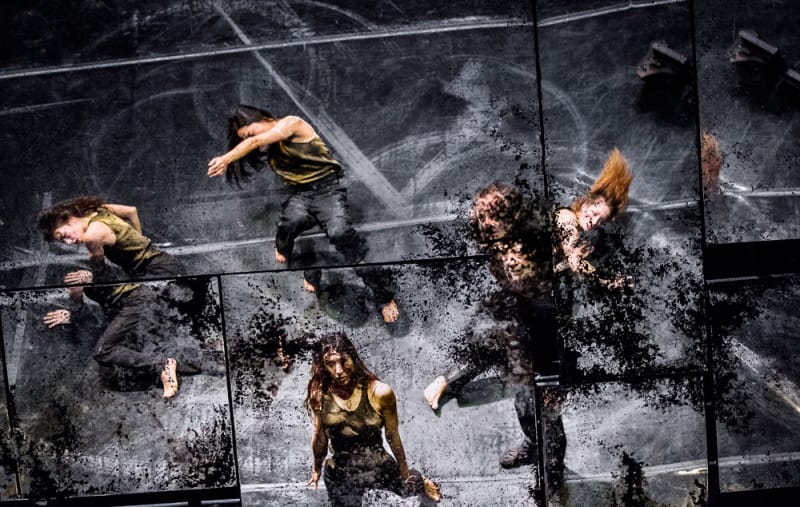 Performers stand on an indoor stage, dressed in a dark trousers and grey shirt and softly lit from above Performer stands on a messy desk on an indoor stage, dressed in a dark trousers and grey shirt and looks at the camera, surrounded by other performers dressed the same and softly lit by golden desk lamps James Thierrée Tabac Rouge Olivier Award