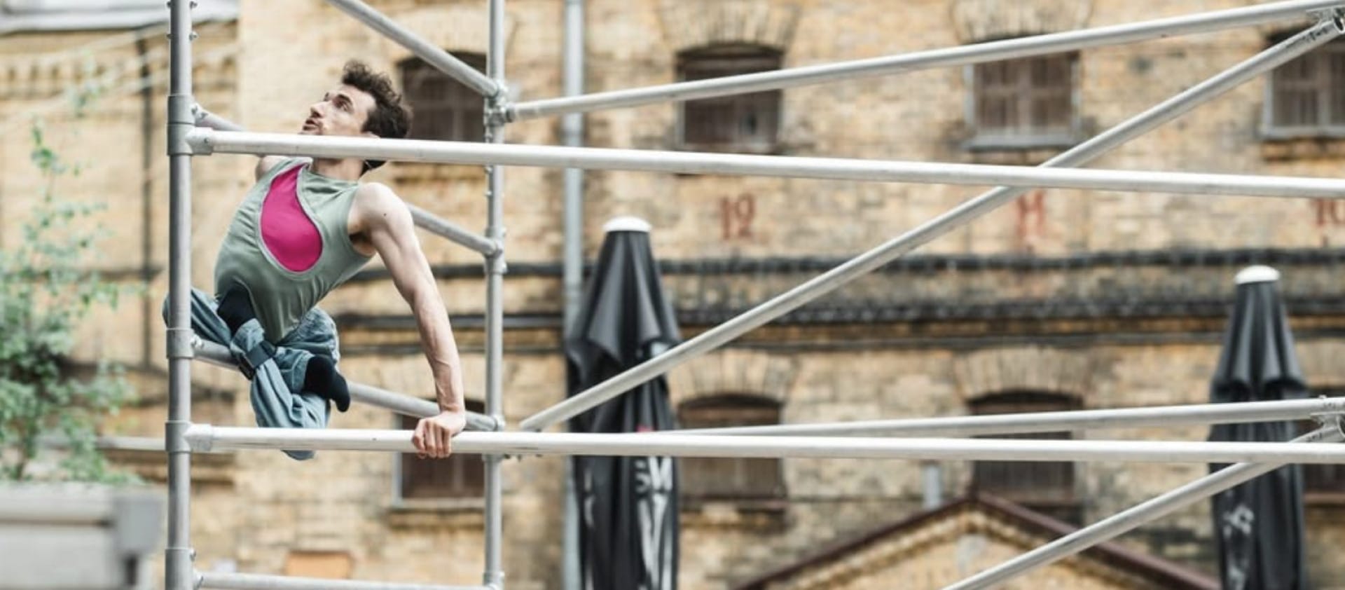 an acrobat with short brown hair wearing a pink and green shirt with blue trousers climbs an outdoor scaffold, their legs are crossed and tied beneath them, an audience watches in front of a brick building Circuliacija Carousel artists thinkers