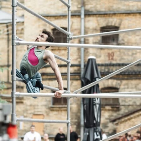 an acrobat with short brown hair wearing a pink and green shirt with blue trousers climbs an outdoor scaffold, their legs are crossed and tied beneath them, an audience watches in front of a brick building Circuliacija Carousel artists thinkers