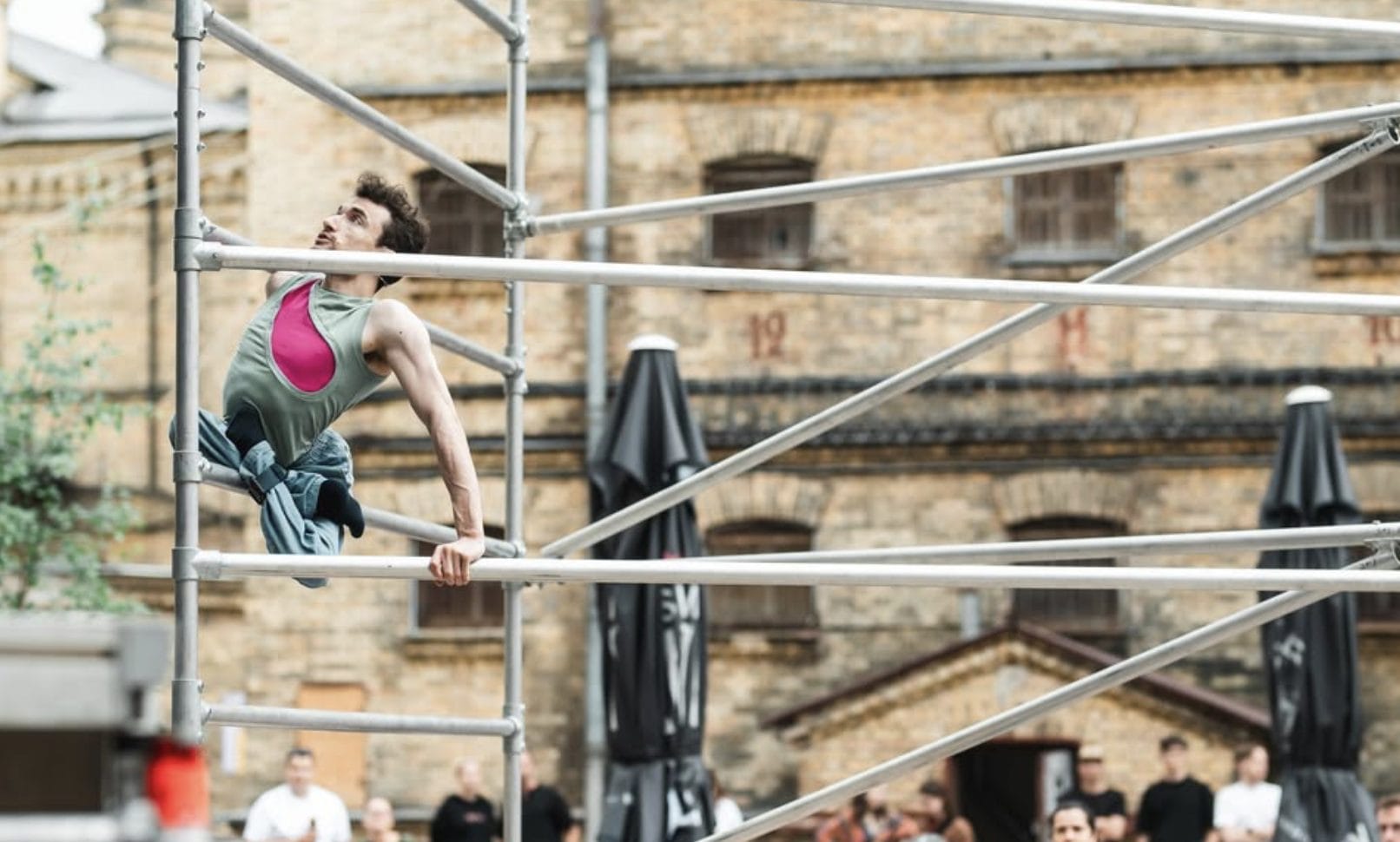 an acrobat with short brown hair wearing a pink and green shirt with blue trousers climbs an outdoor scaffold, their legs are crossed and tied beneath them, an audience watches in front of a brick building Circuliacija Carousel artists thinkers