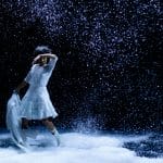 young dancer of colour with curly black hair and a white dress, holding a white coat dances on a darl indoor stage covered in falling fake snow
