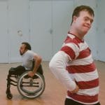 Two individuals in an indoor rehearsal space, one in a wheelchair and another standing with hands on hips, both engaged in movement and expression.