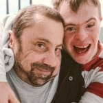 Two dancers from Stopgap Dance Company embrace while looking in to the camera, both have short brown hair, one has a dark beard and is a wheelchair user, they are in an indoor rehearsal room