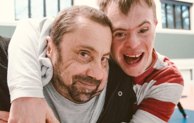 Two dancers from Stopgap Dance Company embrace while looking in to the camera, both have short brown hair, one has a dark beard and is a wheelchair user, they are in an indoor rehearsal room