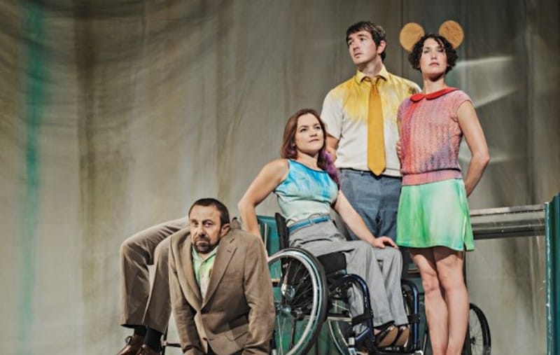 four performers pose for the camera in a indoor rehearsal space, one person who has no lower limbs wears a brown suit and has a dark beard, some 'prop' legs are seen behind them, there is also a wheelchair user with long brown hair wearing a blue vest and grey trousers, the other two are standing up, one wears 'Micky Mouse' ears over curly dark hair, and a green skirt with a red top and the last person has short brown hair and wears grey trousers and a yellow shirt and tie Acrobats residency Stopgap Dance