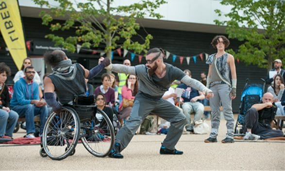 two performers grasp each other's hands in an outdoor festival with an audience looking on, one is a wheelchair user dressed in a grey costume with their hair pulled back, the other dressed similarly wears goggles and has a beard and mohican tied back Greenwich Docklands