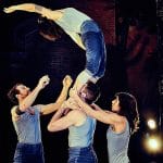 three performers on an indoor stage with a brick wall behind and are spotlit, all wear short blue jeans and pale blue vests, a fourth stands on the shoulders of one and is supported by their legs, as they bend backwards preparing to fall or spring back